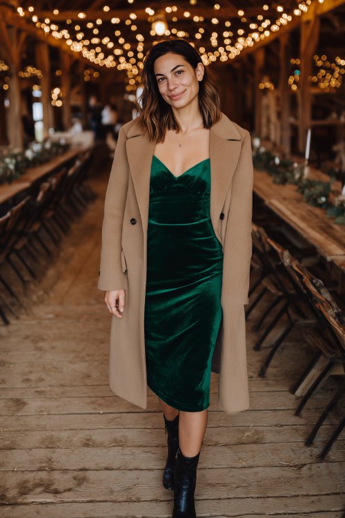 A woman over 40 wearing a hunter green velvet midi dress under a light camel wool coat, styled with black cowboy boots