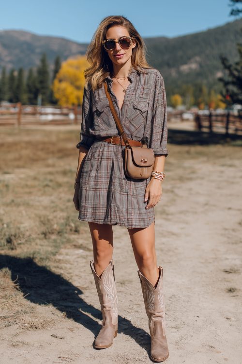 A woman wears a plaid flannel shirt dress with a belt, light brown cowboy boots, and a crossbody bag