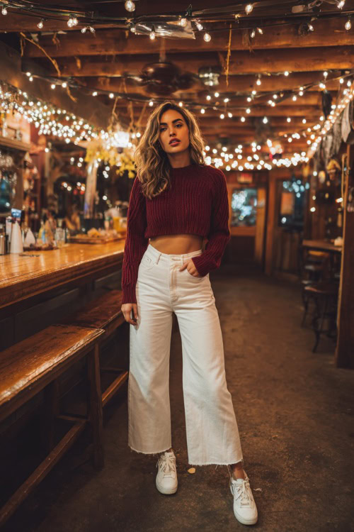 A woman wearing wide-leg jeans, a cropped burgundy knit sweater, and white sneakers, standing at a cozy fall bar with string lights overhead, full body outfit visible