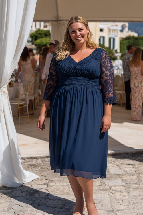 A plus-size woman in a navy chiffon midi dress with lace sleeves and low heels, standing under a white draped pergola at a summer wedding reception
