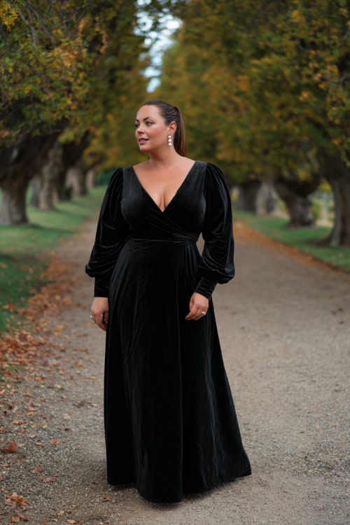 A plus-size woman wears a sleek black velvet gown with long bishop sleeves and a defined waistline, styled with pearl earrings and heels