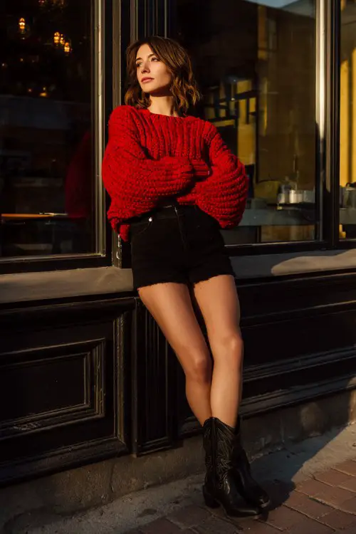 A woman wears a chunky red knit sweater paired with a black mini skirt and cowboy boots