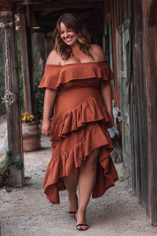 A plus-size woman over 50 in a terracotta off-the-shoulder midi dress with layered ruffles and block heels, walking across a rustic barn wedding path