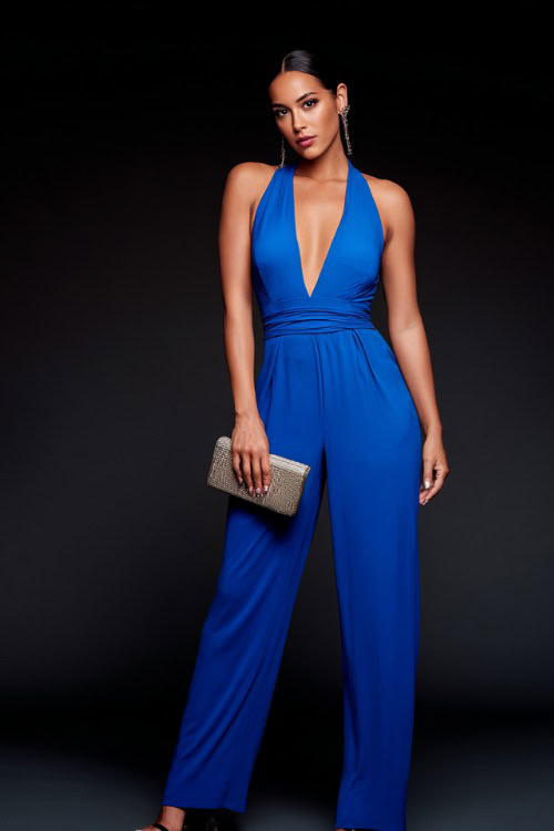 A woman wearing a deep V-neck jumpsuit in bold blue with stiletto heels and a clutch bag, styled for a fashionable summer night out, simple studio background