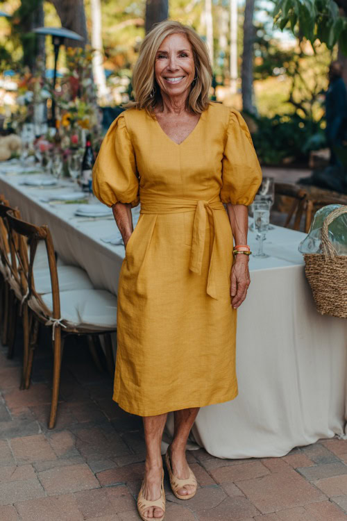 A woman over 50 wearing a mustard yellow belted midi dress with subtle puff sleeves and espadrille wedges, smiling near a boho-chic cocktail wedding setup
