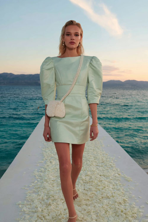 A woman wearing a pastel green cocktail dress with puff sleeves and a pearl-embellished bag, walking along a white path with petals at a sunset ceremony