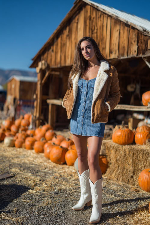 A woman in a denim mini dress paired with a shearling-lined suede jacket and sleek white cowboy boots, standing near a rustic barn decorated with pumpkins