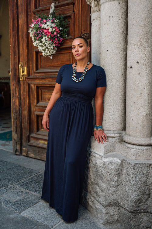 A plus-size woman over 50 wearing a navy blue empire waist maxi dress with short sleeves and a beaded necklace, standing near a classic church entrance with floral decorations