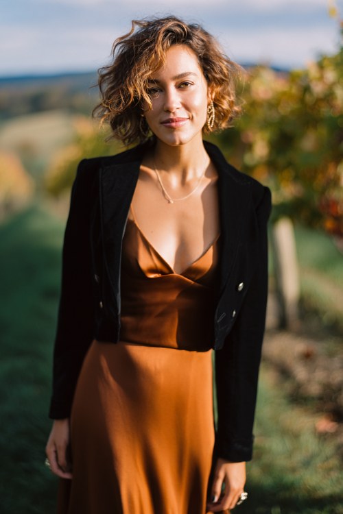 A full body image of woman wears a copper silk slip gown with a flowing hem, layered under a black velvet cropped jacket