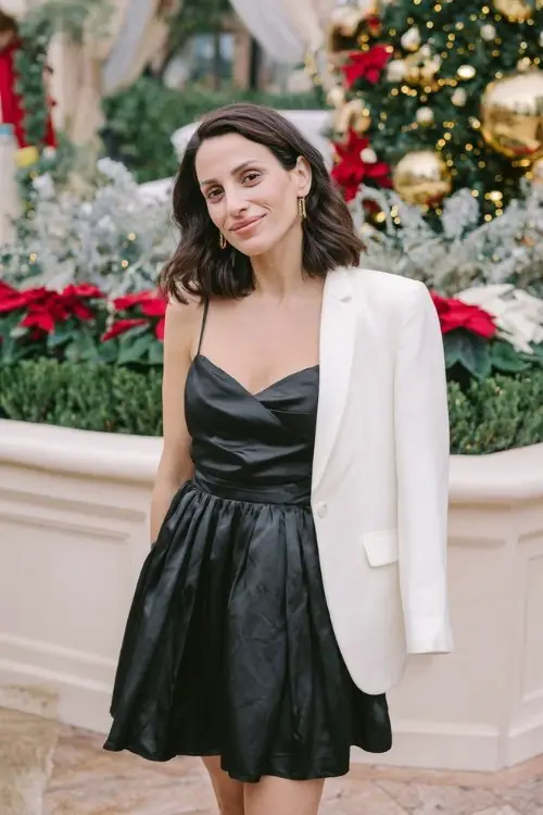 A woman over 30 wears a black satin cocktail dress with a pleated skirt and a tailored white blazer draped over her shoulders