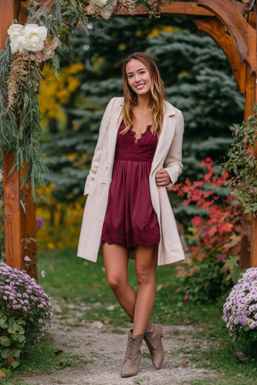 A woman wears a maroon A-line dress with a flowy skirt and lace sleeves, styled with a light tan wrap coat and ankle boots, posing under a decorated wedding arch in a fall garden setting