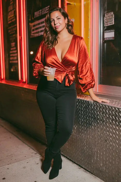 A curvy woman wears a metallic red wrap top, skinny black jeans, and pointed-toe ankle boots, holding a drink