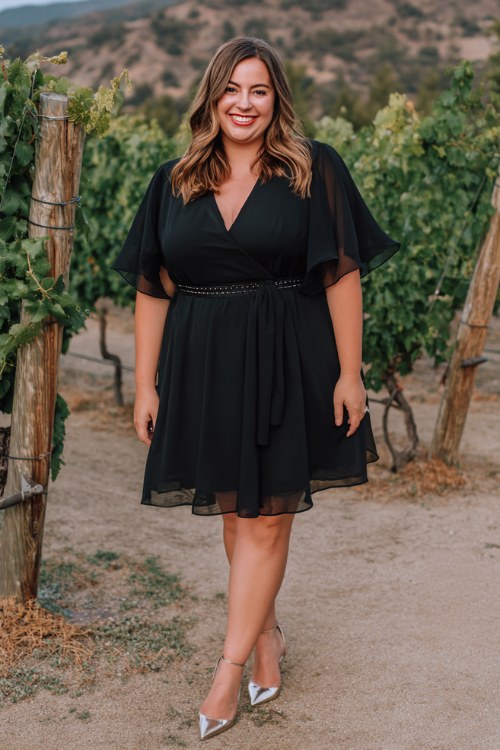 A plus-size woman wears a black A-line chiffon dress with layered flutter sleeves and a belted waist