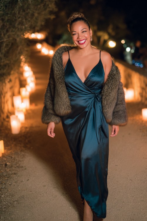 A curvy woman wears a dark teal satin gown with a wrap silhouette and faux-fur shawl