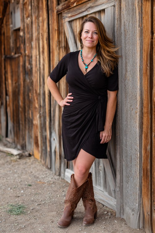 A curvy woman wears a black wrap dress with brown cowboy boots and turquoise jewelry