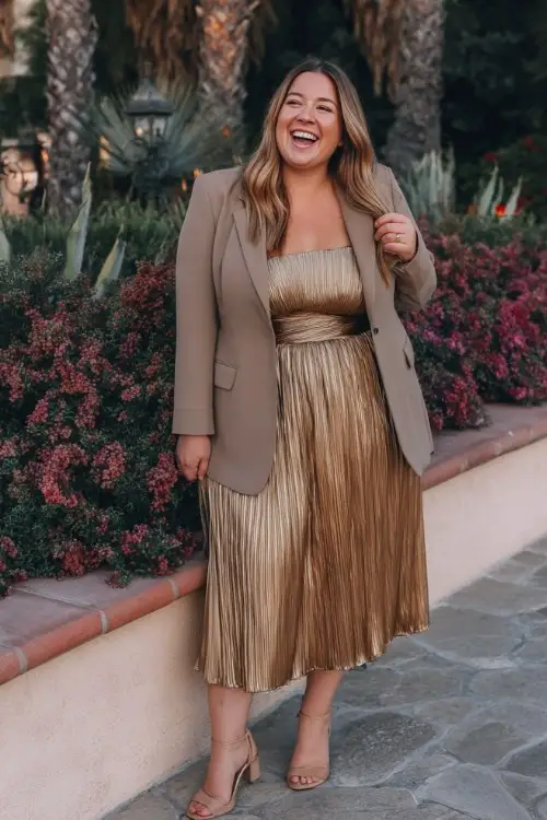 A plus-size woman wears a bronze metallic midi dress with a subtle pleated finish, paired with a structured blazer and patent heels