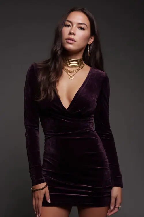 A woman in a deep plum velvet mini dress with long sleeves and gold accessories, styled for a luxe and moody summer night out, simple studio background