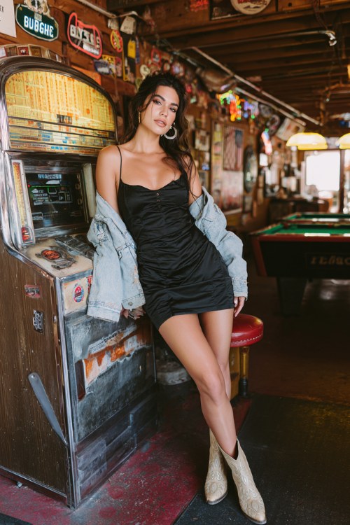 A woman wears a black slip dress layered under a cropped denim jacket with western booties