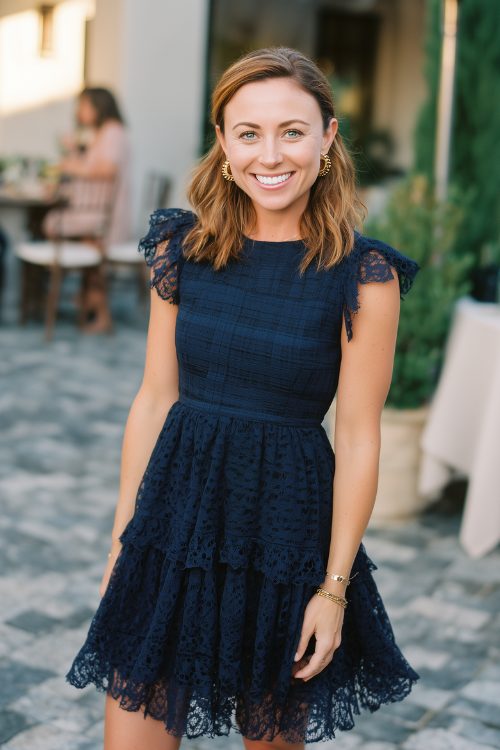 A petite woman wears a navy fit-and-flare dress with tiered lace and a subtle plaid overlay