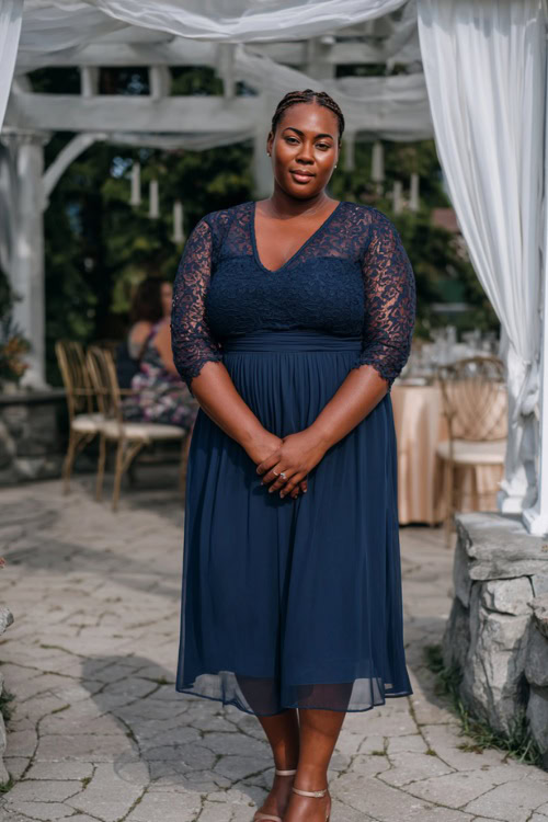A plus-size woman in a navy chiffon midi dress with lace sleeves and low heels, standing under a white draped pergola at a summer wedding reception