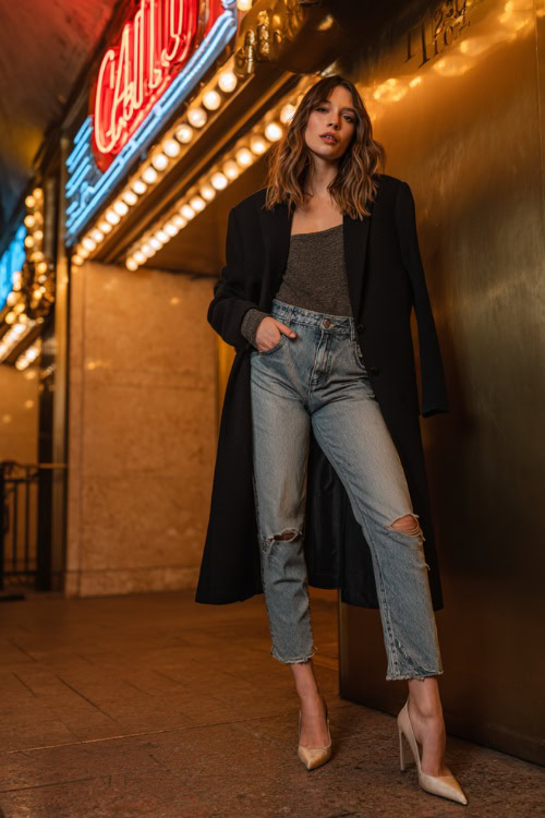 A woman wearing skinny jeans, an oversized blazer, and pointed heels, standing outside a trendy fall club entrance with glowing signs, full body outfit visible