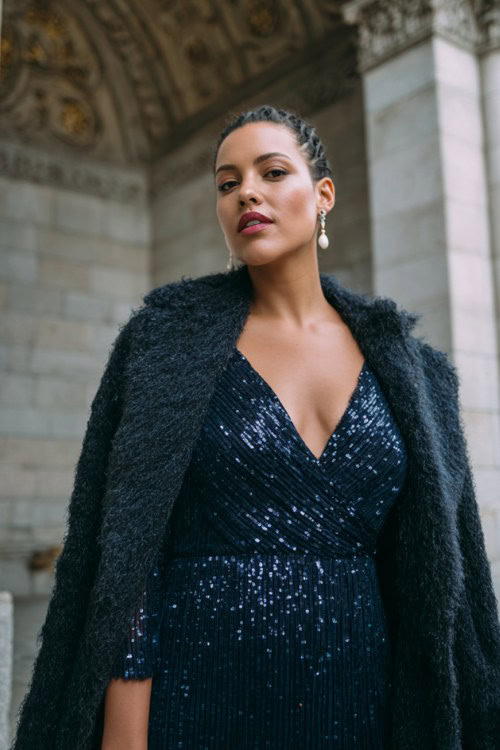 A curvy woman wears a navy sequin evening gown with long sleeves, styled with a dark wool wrap coat and pearl earrings