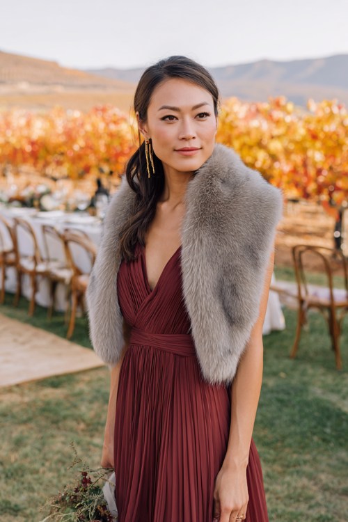 A woman wears a maroon pleated maxi dress styled with a light gray fur stole, standing by a vineyard ceremony with orange and gold leaves in the background