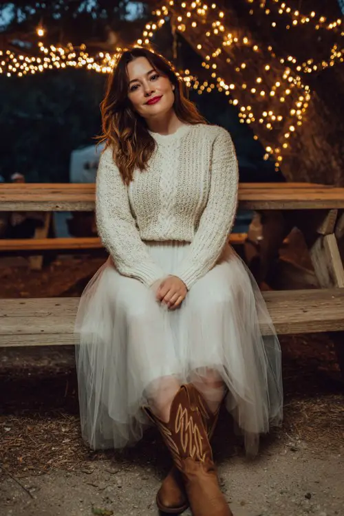 A curvy woman wears brown cowboy boots with a cream cable-knit sweater and a tulle skirt