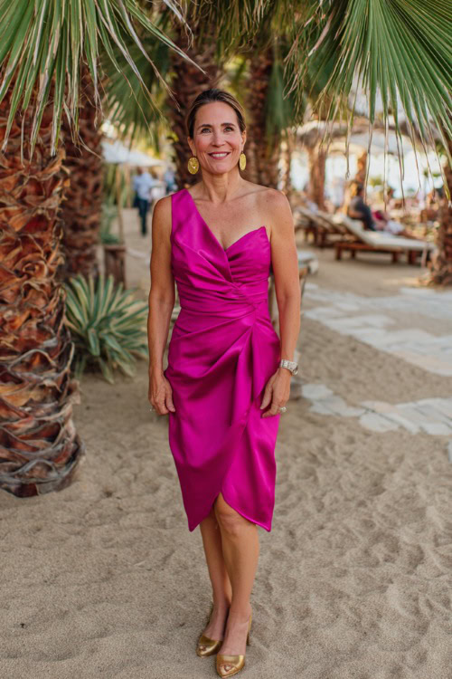 A woman over 50 wearing a bold fuchsia cocktail dress with asymmetrical neckline and gold heels