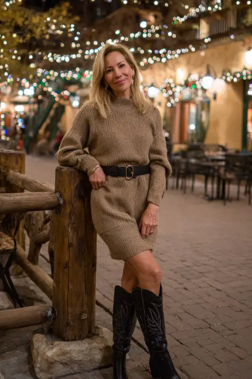 A woman over 40 wears a camel knit sweater dress with a belt at the waist and tall black cowboy boots