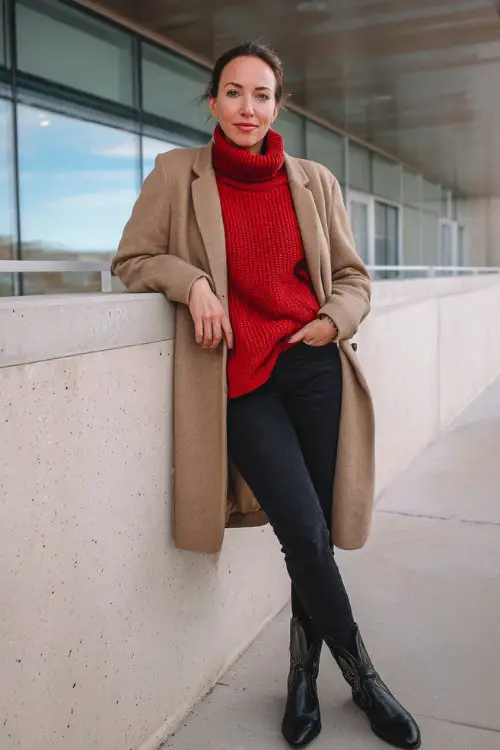 A woman over 40 wears a red turtleneck sweater layered with a camel coat, black skinny jeans, and black cowboy boots