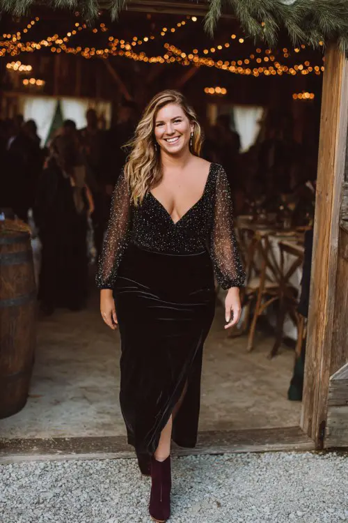 A curvy woman wears a black sequin cocktail dress with sheer mesh sleeves and dark plum velvet boots