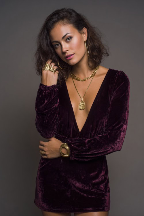 A woman in a deep plum velvet mini dress with long sleeves and gold accessories, styled for a luxe and moody summer night out, simple studio background