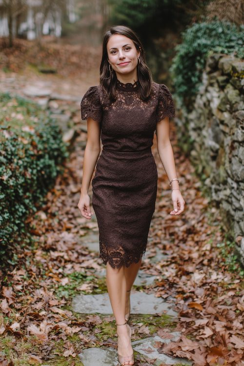 A petite woman in a chocolate brown lace dress with scalloped sleeves and simple jewelry, walking through a leaf-covered stone path