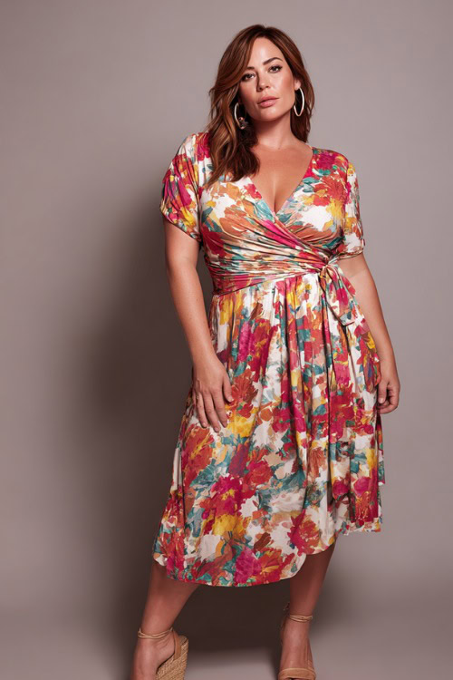 A plus-size woman wearing a wrap midi dress in bold floral print with wedge sandals and hoop earrings, styled for a summer evening out, simple studio background