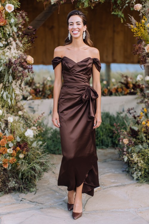 A woman wears a chocolate brown satin gown with an off-shoulder neckline and cascading side ruffle