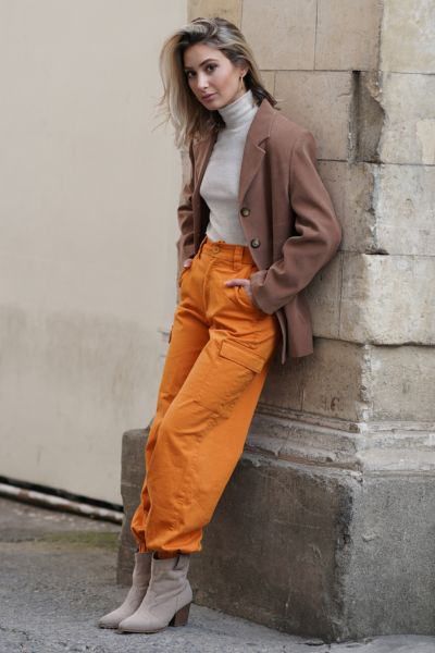 A woman wears short cowboy boots with orange cargo pants, turtleneck and coat
