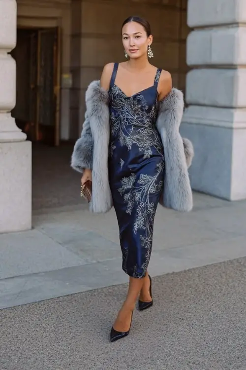 woman wears a navy dress with silver sequin overlay in a cascading vine pattern, styled with a cropped grey fur coat