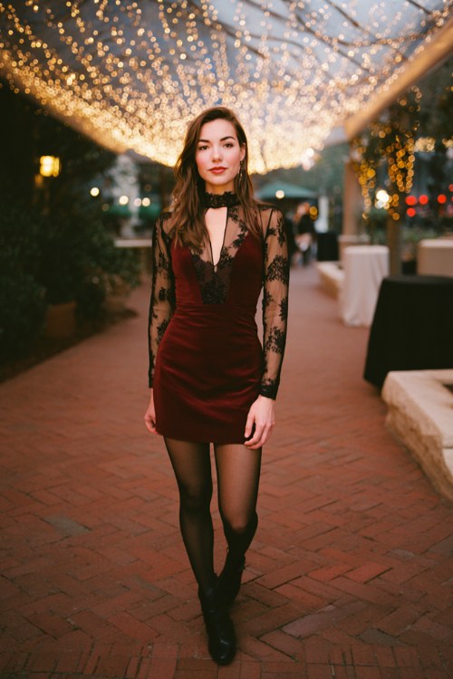 A woman over 30 wears a dark red fitted cocktail dress with a sheer embroidered overlay, black tights, and glossy ankle boots