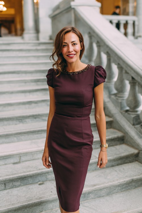 A woman over 50 wears a plum-colored sheath dress with structured short sleeves and subtle beading at the neckline