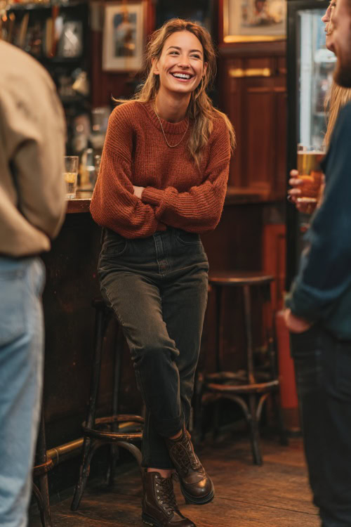 A woman in dark straight-leg jeans, a rust-colored knit sweater, and Chelsea boots, laughing with friends at a casual fall bar