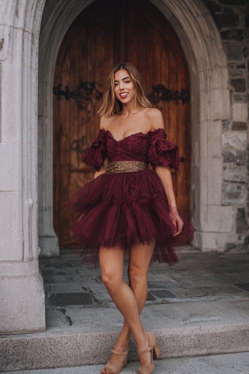 A woman wears a burgundy off-shoulder cocktail dress with tulle layers and a crochet bodice, finished with suede block heels and a leather braided belt