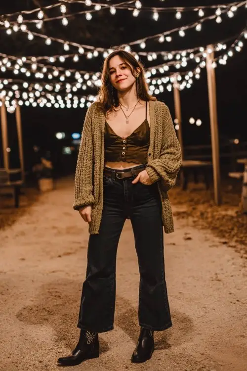 A woman wears flared jeans with black cowboy boots outfit for fall, layered with a forest green knit cardigan over a satin camisole
