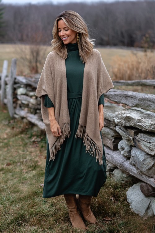 A woman wears a forest green A-line midi dress with mesh sleeves, layered under a camel knit poncho