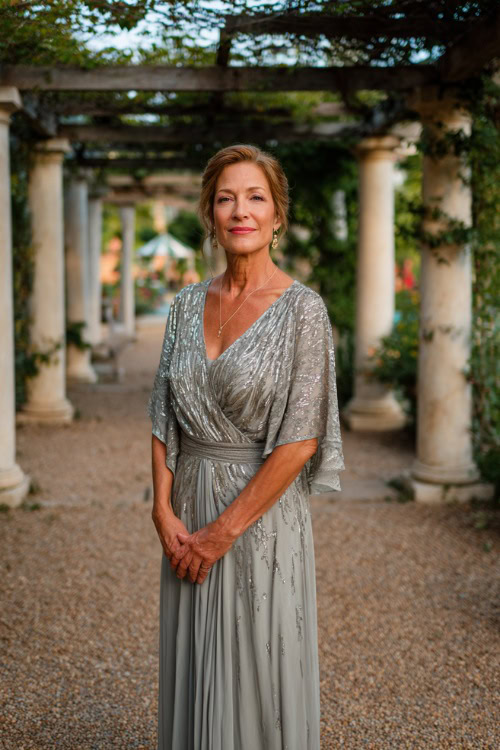 A woman over 50 in a soft gray sequin-embellished gown with flutter sleeves and a flowing skirt, standing under a decorated pergola at a sunset garden wedding