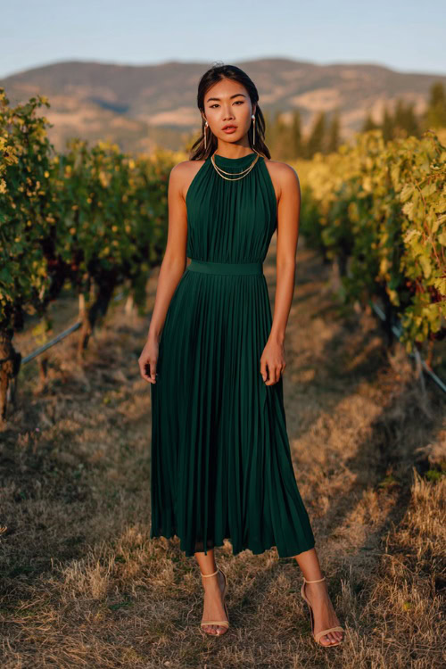 A woman wears a forest green pleated chiffon midi dress with a fitted waist and flowy skirt, accessorized with ankle strap heels