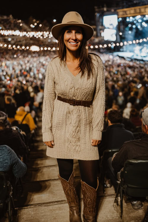 A woman over 40 wears a long sleeve knit sweater dress with a belted waist, black tights, and tall cowboy boots