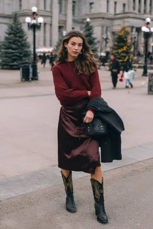 A woman wears black cowboy boots with a burgundy knit sweater and a satin slip skirt, holding her coat over her arm