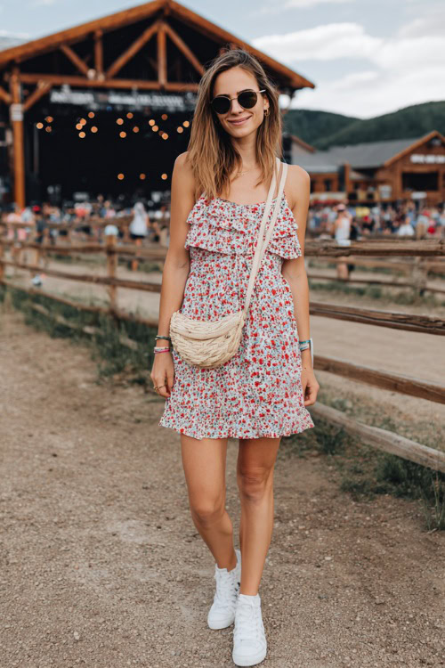 A woman wearing a smocked floral sundress with ruffle straps and white canvas sneakers, accessorized with a straw crossbody bag and sunglasses