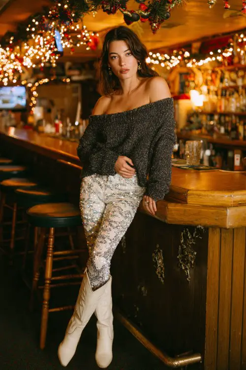 A woman wears sparkly leggings with a chunky knit off-the-shoulder sweater and white cowboy boots
